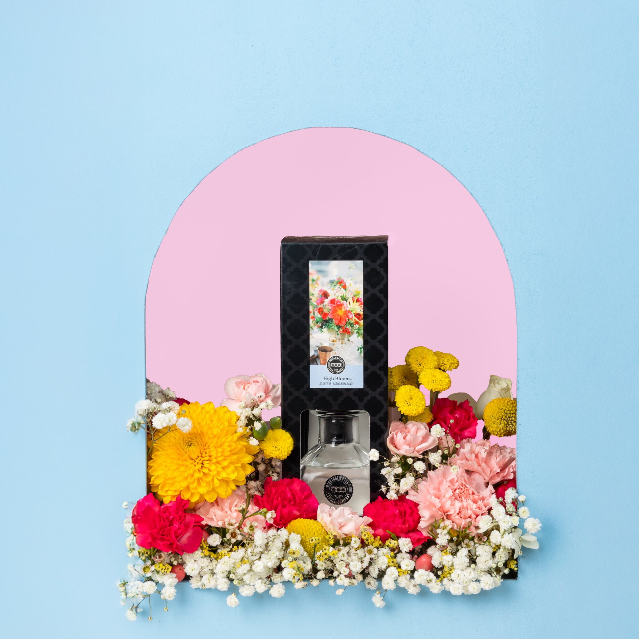 The High Bloom Reed Diffuser by Bridgewater Candle is showcased in an arched alcove with a pink background, accented by colorful flowers against a light blue wall for a charming home décor and premium fragrance experience.