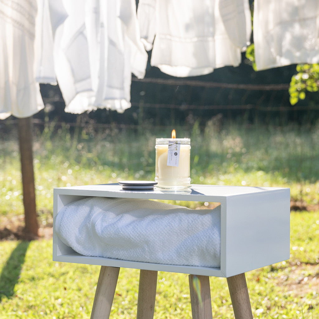 The Laundry Line 8oz Jar Candle by Bridgewater Candle Company glows in a glass jar on a white shelf with a towel outdoors, while fresh white laundry sways on the line in the sunny, grassy background.