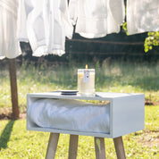 The Laundry Line 8oz Jar Candle by Bridgewater Candle Company glows in a glass jar on a white shelf with a towel outdoors, while fresh white laundry sways on the line in the sunny, grassy background.