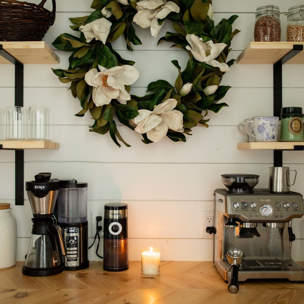 A coffee station with a coffee maker, grinder, espresso machine, and a glowing Bridgewater Candle Company Sweet Magnolia 18oz Large Jar Candle on the wooden counter. Cups and jars are on shelves above; a white floral wreath hangs on the shiplap wall.