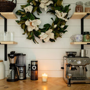 A coffee station with a coffee maker, grinder, espresso machine, and a glowing Bridgewater Candle Company Sweet Magnolia 18oz Large Jar Candle on the wooden counter. Cups and jars are on shelves above; a white floral wreath hangs on the shiplap wall.