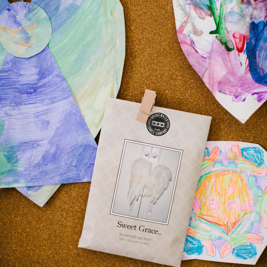 A Bridgewater Candle Company Sweet Grace Sachet with angel wings is clipped to a corkboard, surrounded by colorful, heart-shaped childrens drawings, adding a delightful home fragrance.