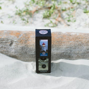 The Nantucket Coast Reed Diffuser, featuring a lighthouse image on its label, stands upright in sand before a weathered log, with green plants and blurred sand in the background.