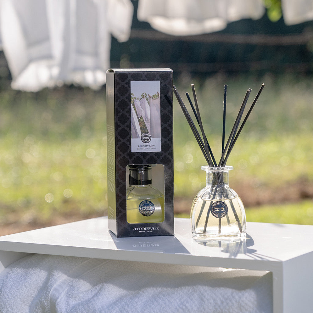 A clear Bridgewater Candle Company Laundry Line Reed Diffuser with black fiber reeds is displayed beside its box outdoors on a white surface, with sunlight and white laundry in the background, suggesting a fresh laundry scent.