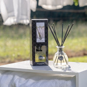 A clear Bridgewater Candle Company Laundry Line Reed Diffuser with black fiber reeds is displayed beside its box outdoors on a white surface, with sunlight and white laundry in the background, suggesting a fresh laundry scent.