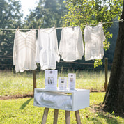 The Laundry Line Flameless Bundle by Bridgewater Candle Company sits on a stool outdoors with white blouses drying on a clothesline nearby and trees and grass in the sunny background.