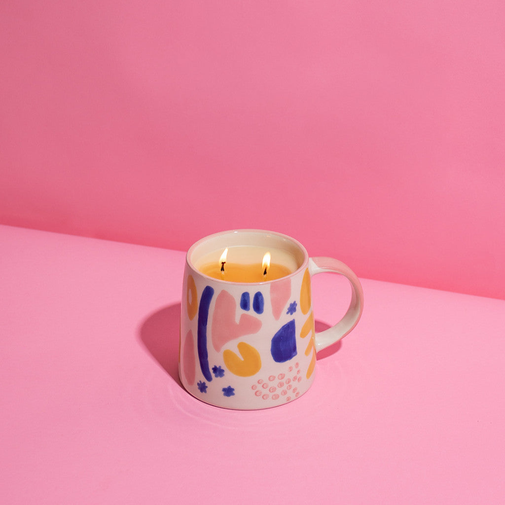 A ceramic mug with a colorful abstract design holds a lit Bridgewater Candle Company Sweet Grace Collection Candle #047, made from soy-blend wax for a clean burn, on a pink surface against a pink background.