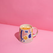 A ceramic mug with a colorful abstract design holds a lit Bridgewater Candle Company Sweet Grace Collection Candle #047, made from soy-blend wax for a clean burn, on a pink surface against a pink background.