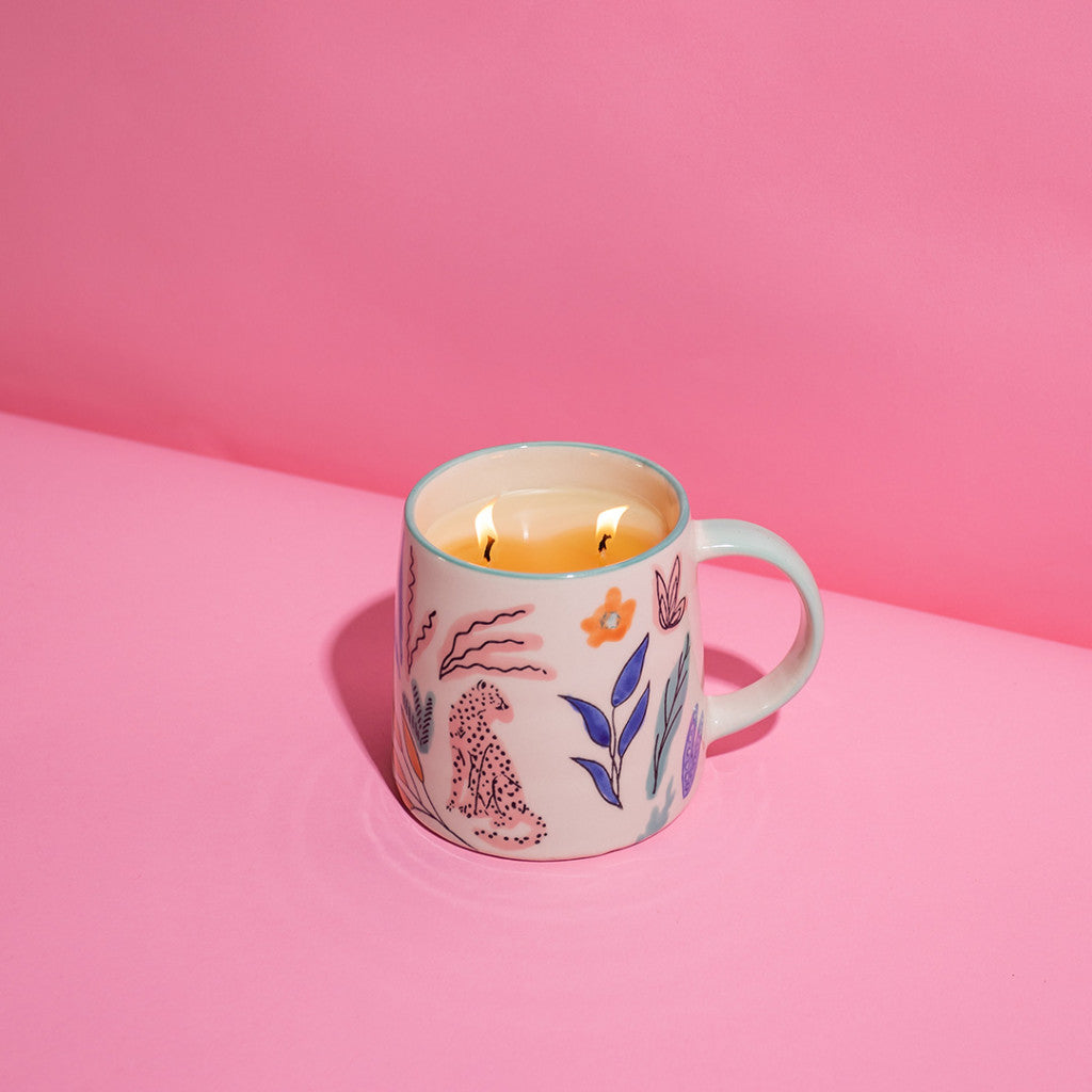 The Bridgewater Candle Company Sweet Grace Collection Candle #048, in a decorative ceramic mug featuring jeweled jungle motifs and vibrant florals, sits on a smooth pink surface with a coordinating pink background.