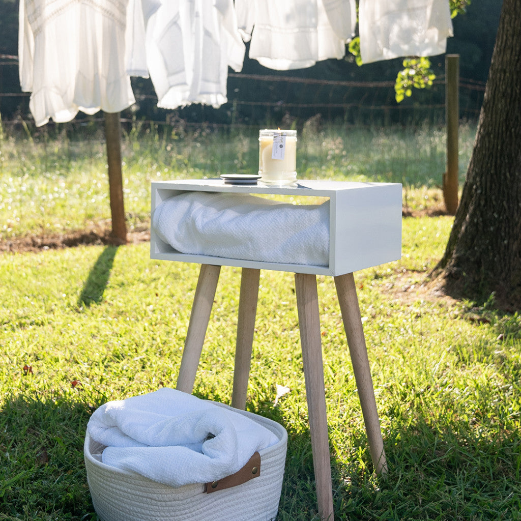 A small white table with wooden legs holds a folded towel, a Bridgewater Candle Company Laundry Line 8oz Jar Candle, and a plate. Nearby on the grass, a basket with a towel sits as sunlit laundry hangs fresh in the background.