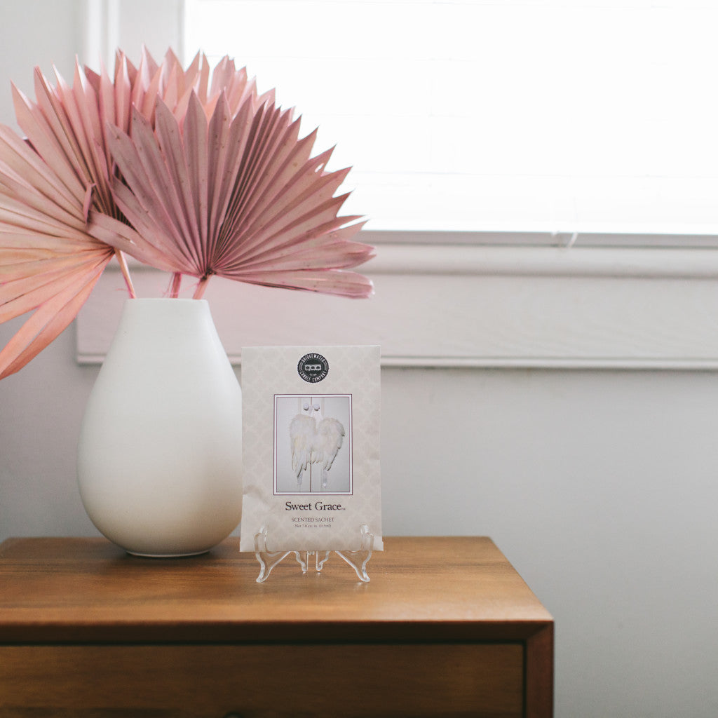 A white vase with pink dried palm leaves sits on a wooden table beside a Sweet Grace Sachet by Bridgewater Candle Company and a card on a stand, set against a white wall and window, adding elegance and charm to the room.