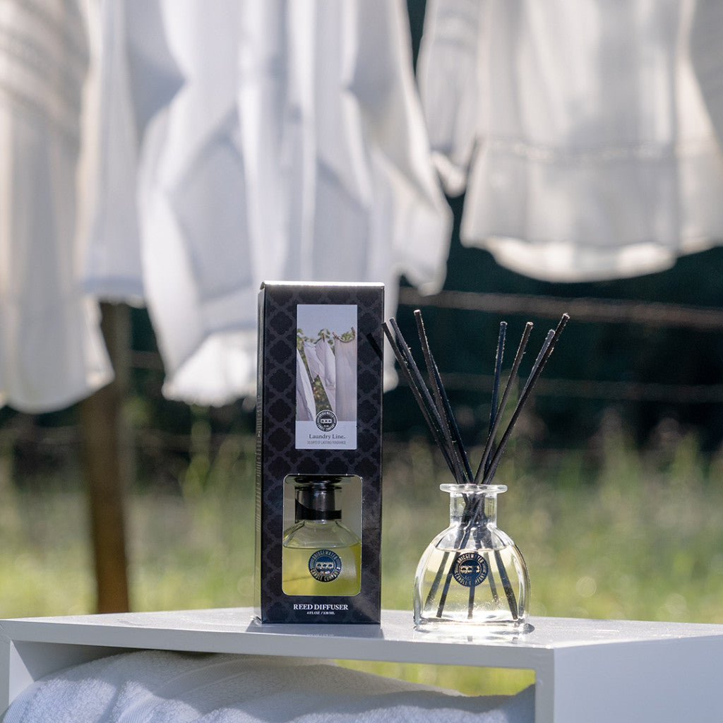 A Bridgewater Candle Company Laundry Line Reed Diffuser and its box rest on a sunlit white surface outdoors, with white fabric in the background suggesting a fresh laundry scent. Fiber reeds add to the serene, airy ambiance.