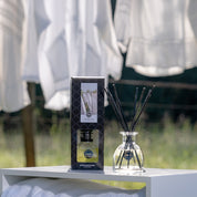 A Bridgewater Candle Company Laundry Line Reed Diffuser and its box rest on a sunlit white surface outdoors, with white fabric in the background suggesting a fresh laundry scent. Fiber reeds add to the serene, airy ambiance.