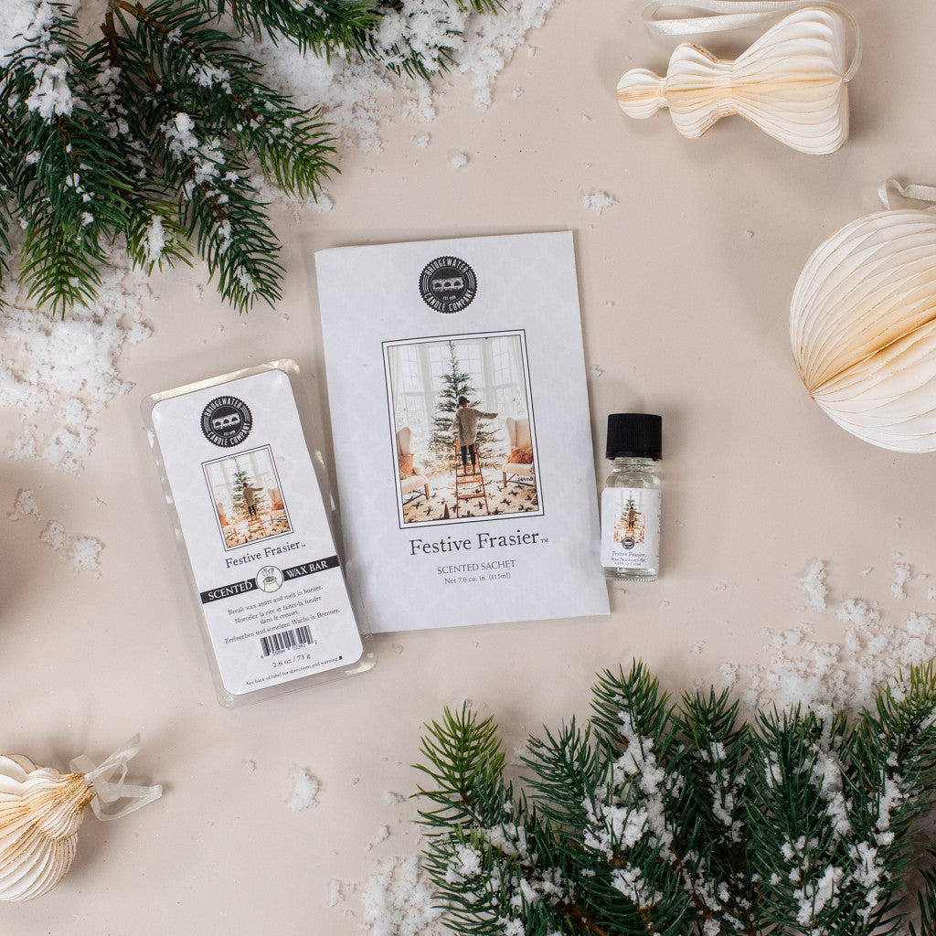 Flat lay of Bridgewater Candle Companys Festive Frasier Flameless Bundle, featuring home fragrance oil, pine branches, artificial snow, and cream-colored paper ornaments on a light surface.
