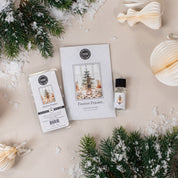 Flat lay of Bridgewater Candle Companys Festive Frasier Flameless Bundle, featuring home fragrance oil, pine branches, artificial snow, and cream-colored paper ornaments on a light surface.