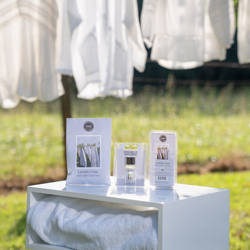 The Laundry Line Flameless Bundle by Bridgewater Candle Company sits outdoors atop a white cabinet with folded towel and laundry items. Behind it, white clothes hang on a line, surrounded by greenery and sunlight.