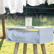 The Bridgewater Candle Company Laundry Line 8oz Jar Candle sits on a white box with a towel, placed on wooden legs outdoors as laundry dries in the sun, capturing a fresh, clean scent inspired by lines of sun-warmed linens.