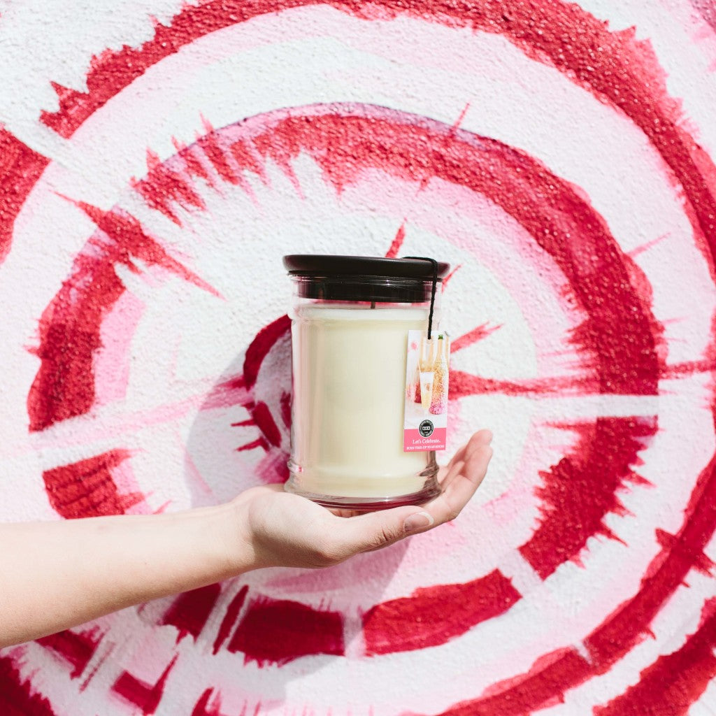 A hand holds a Bridgewater Candle Company Lets Celebrate 18oz Large Jar Candle with a black lid and pink tag, set against a red and white spiral mural painted on a textured wall.
