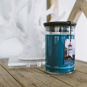 A Bridgewater Candle Company Nantucket Coast 18oz Large Jar Candle with a black lid rests on a wooden table. A tag featuring a lighthouse hints at its refreshing scent, while white decorative coral accents the background.