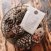 A wire basket of pinecones holds a Bridgewater Candle Company Sweet Grace Sachet, featuring an angel wings illustration, resting on a rustic wooden surface.