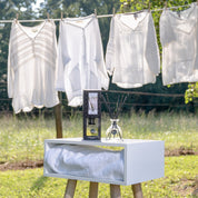 A white box with a blanket, the Bridgewater Candle Company Laundry Line Reed Diffuser, and a fragrance bottle sits outdoors on wooden legs, with white blouses on a clothesline and green trees in the background.
