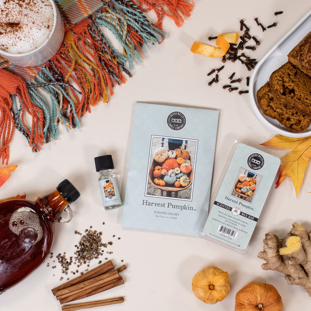 Flat lay of autumn items featuring the Bridgewater Candle Company Harvest Pumpkin Flameless Bundle, a cozy drink, pumpkin bread, spices, syrup, ginger, and a plaid scarf arranged on a cream background.