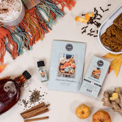 Flat lay of autumn items featuring the Bridgewater Candle Company Harvest Pumpkin Flameless Bundle, a cozy drink, pumpkin bread, spices, syrup, ginger, and a plaid scarf arranged on a cream background.