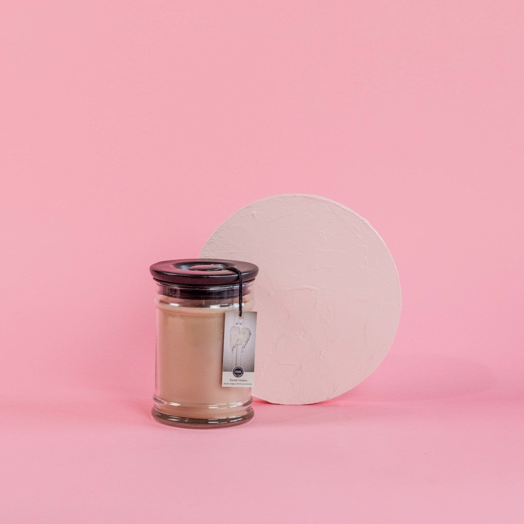 The Bridgewater Candle Company’s Sweet Grace 8oz Jar Candle, in a beige glass jar with a black lid and tag, sits beside a round white textured disc on a pink background—its scent blends sparkling tea with hints of passion fruit.