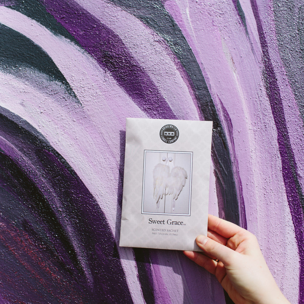 A hand holds a Bridgewater Candle Company Sweet Grace Sachet with angel wings on the packaging, set against a purple and black abstract painted wall.