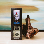 A Nantucket Coast Reed Diffuser in its box sits on a wooden surface next to driftwood, with fiber reeds and a blurred painting of water and sky in the background.