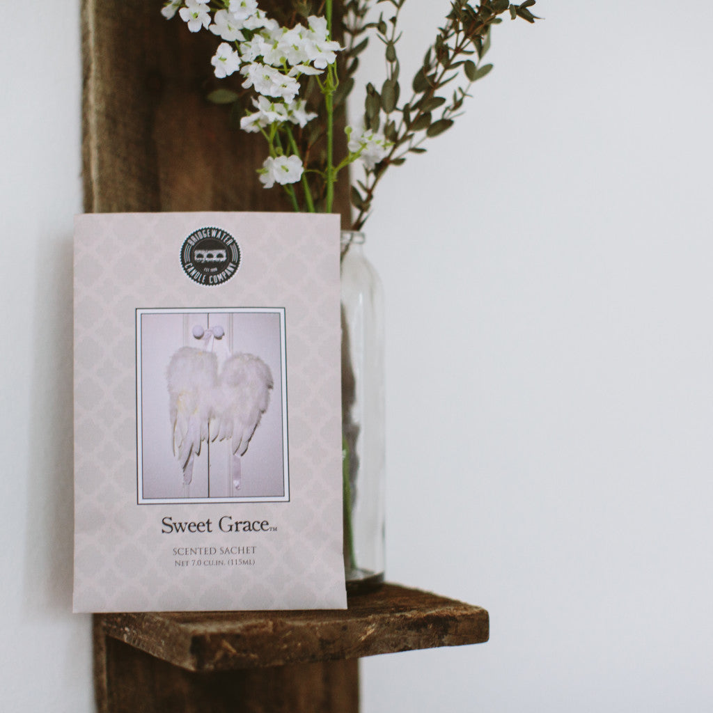 A Bridgewater Candle Company Sweet Grace Sachet, adorned with white angel wings, sits on a wooden shelf beside a glass vase of white flowers, bringing delightful fragrance to the room.