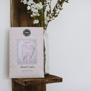 A Bridgewater Candle Company Sweet Grace Sachet, adorned with white angel wings, sits on a wooden shelf beside a glass vase of white flowers, bringing delightful fragrance to the room.