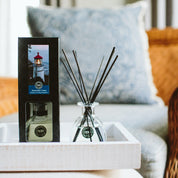 A Nantucket Coast Reed Diffuser with fiber reeds and its box sit on a white tray atop a light surface, while a patterned cushion and cozy armchair blur softly in the background.