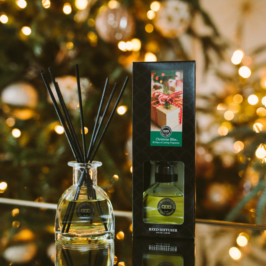 A Bridgewater Candle Company Christmas Bliss Reed Diffuser, with its box, rests on a reflective surface before a glowing Christmas tree—an ideal festive home décor accent.