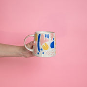 A hand with pink nail polish holds a ceramic mug featuring an abstract blue, yellow, and pink design against a pink background—perfect for enjoying the clean burn of your Sweet Grace Collection Candle #047 by Bridgewater Candle Company.